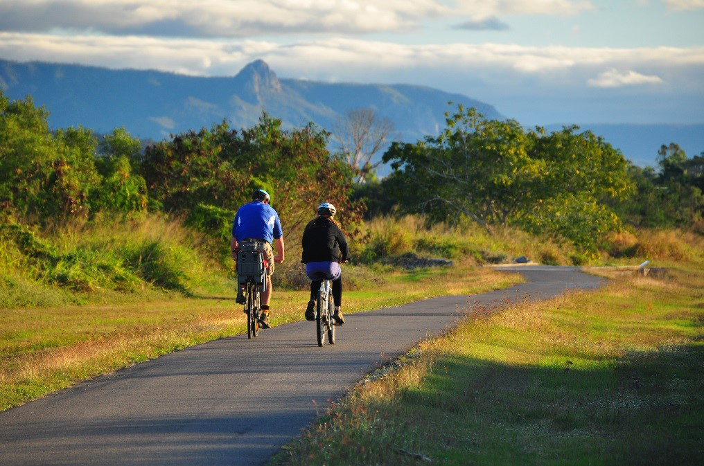 Two riders riding on a bike path in the foreground of mountains.