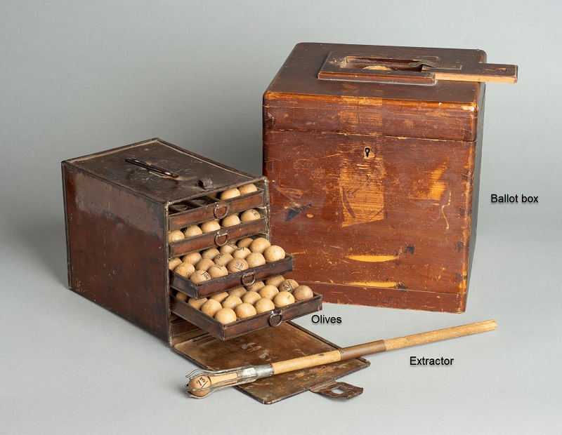 A timber ballot box, timber olives (marbles) encased in a metal box, and timber extractor tool with pronged end holding an olive.