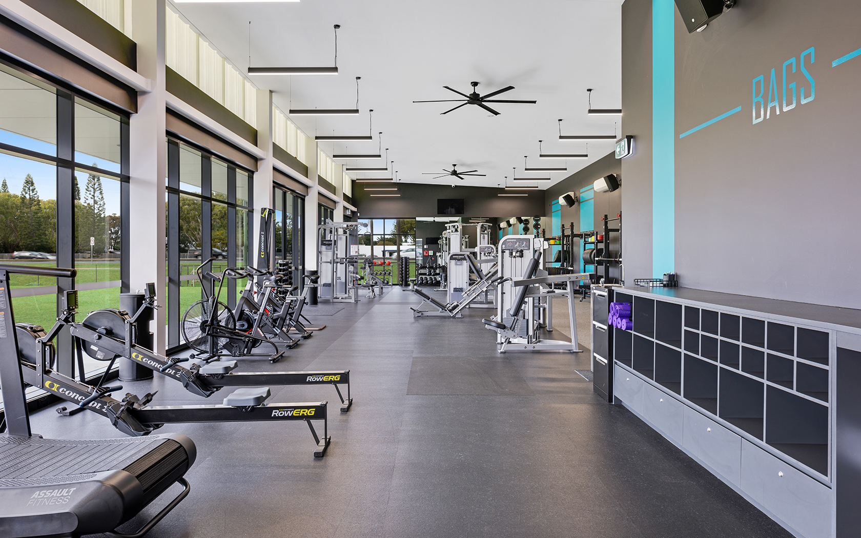 Interior of a gym with exercise equipment and large windows looking out to a green space.
