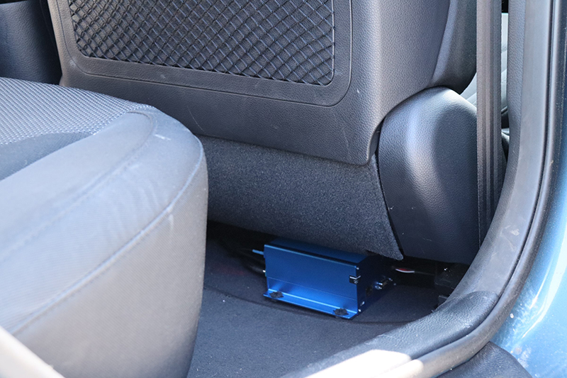 Photo of the inside of a car behind the driver's seat showing a small blue box affixed to the floor under the seat.