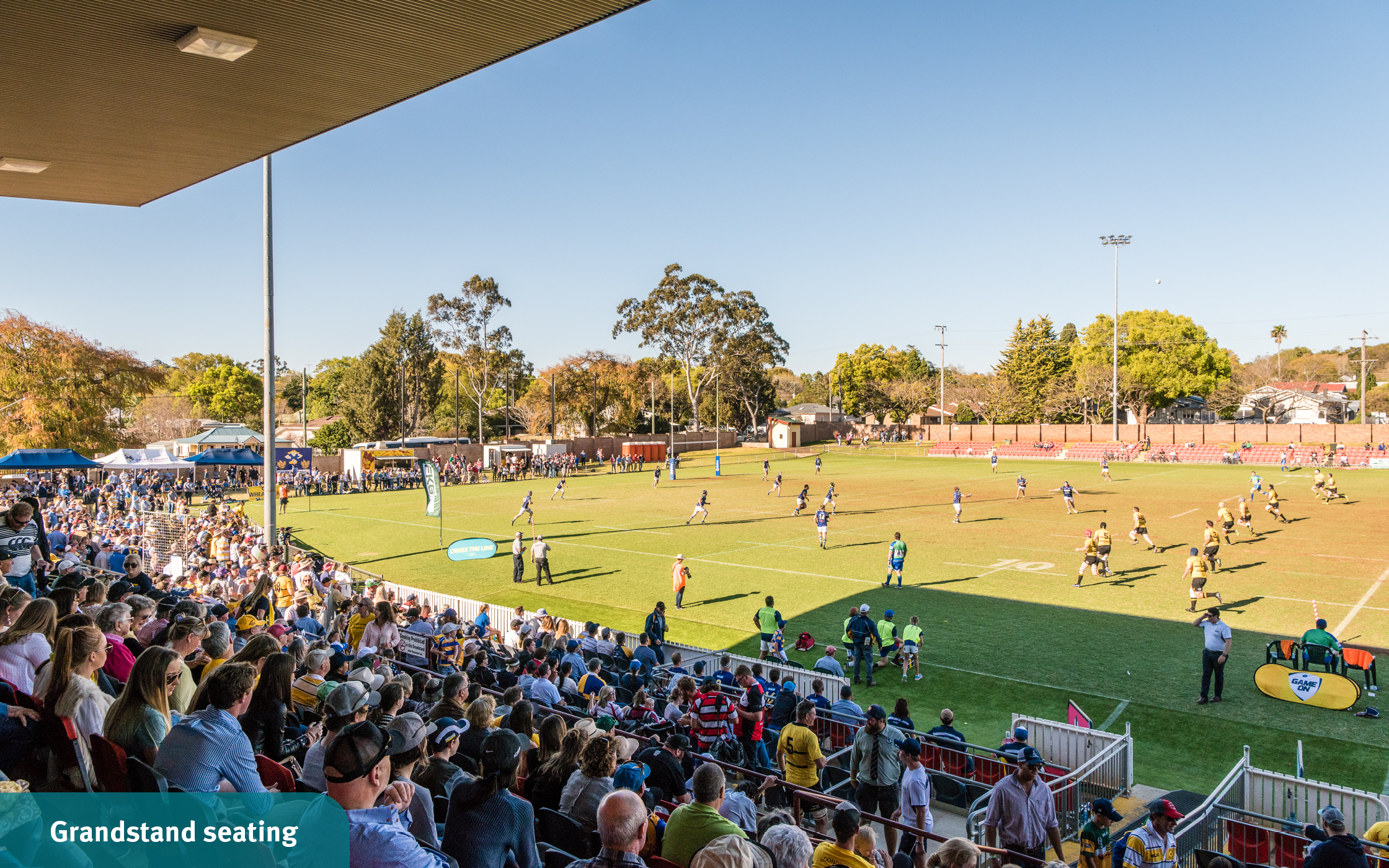 Spectators watch football players running across the field from the cover of the grandstand at Toowoomba Sports Grounds.