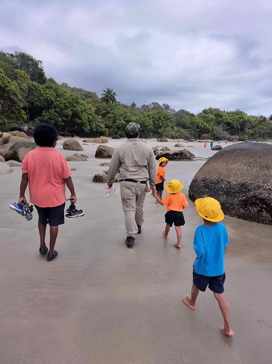 Photo of Gunggandji-Mandingalbay Yidinji ranger and teacher walking along the beach with children for ‘Kindy on Country’ for the Yarrabah School.