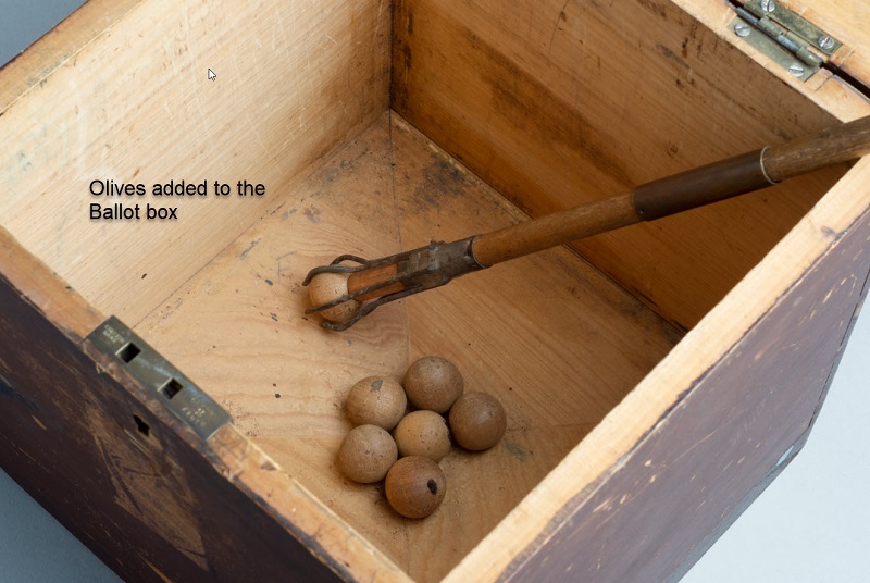 A view inside the ballot box. Six timber olives (marbles) are inside the box and a timber extractor tool with pronged end holds an olive.