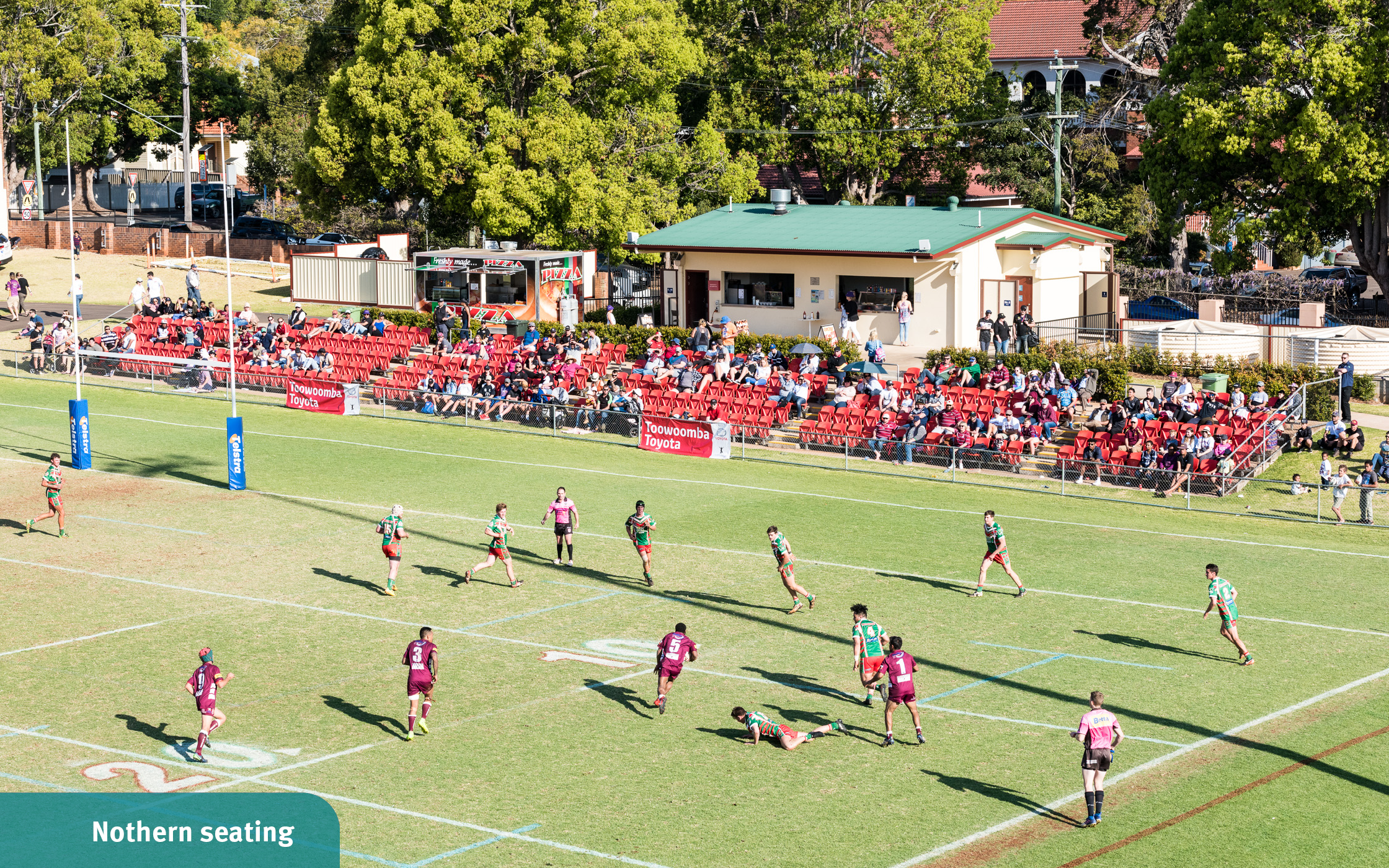 Spectators sitting in red outdoor seating as they watch from the northern end of the field as football players run across the field at Toowoomba Sports Grounds.