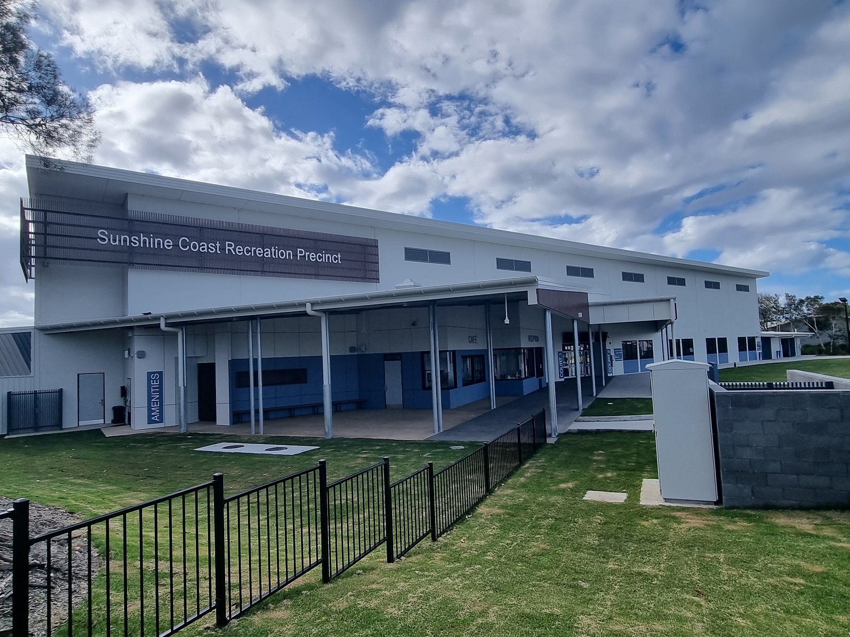 Outdoor view of the fenced Sunshine Coast Recreation Precinct with cafe, reception, and amenities signs, featuring grassed areas and paved spaces under a roofed area