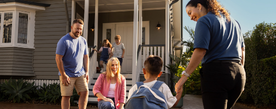 Small child holding the hand of middle aged female whilst a middle aged couple stand and kneel in front of the child. Two young children stand on the stairs behind the adults. All are in front of a grey wooden house.