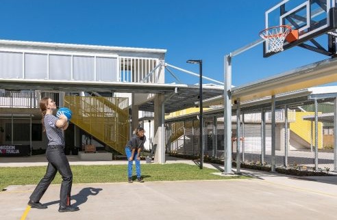 A person is standing on a basketball court and is preparing to throw the basketball into the hoop. Another person is at the side of the basketball court with their hands on their knees.
