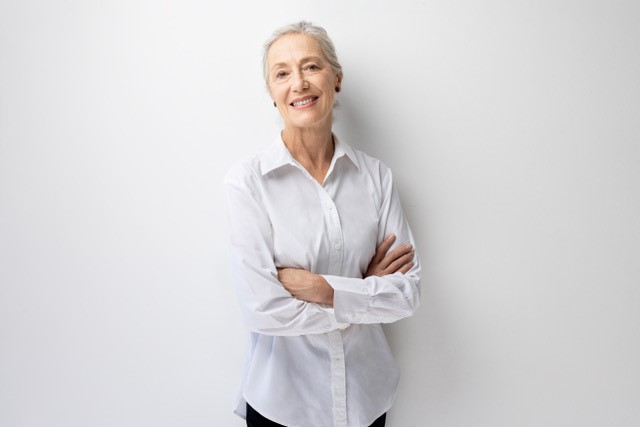 Shane Carroll has fair skin, white hair pulled off their face, wearing a white collared shirt, black pants facing the camera with their arms crossed. with a white background