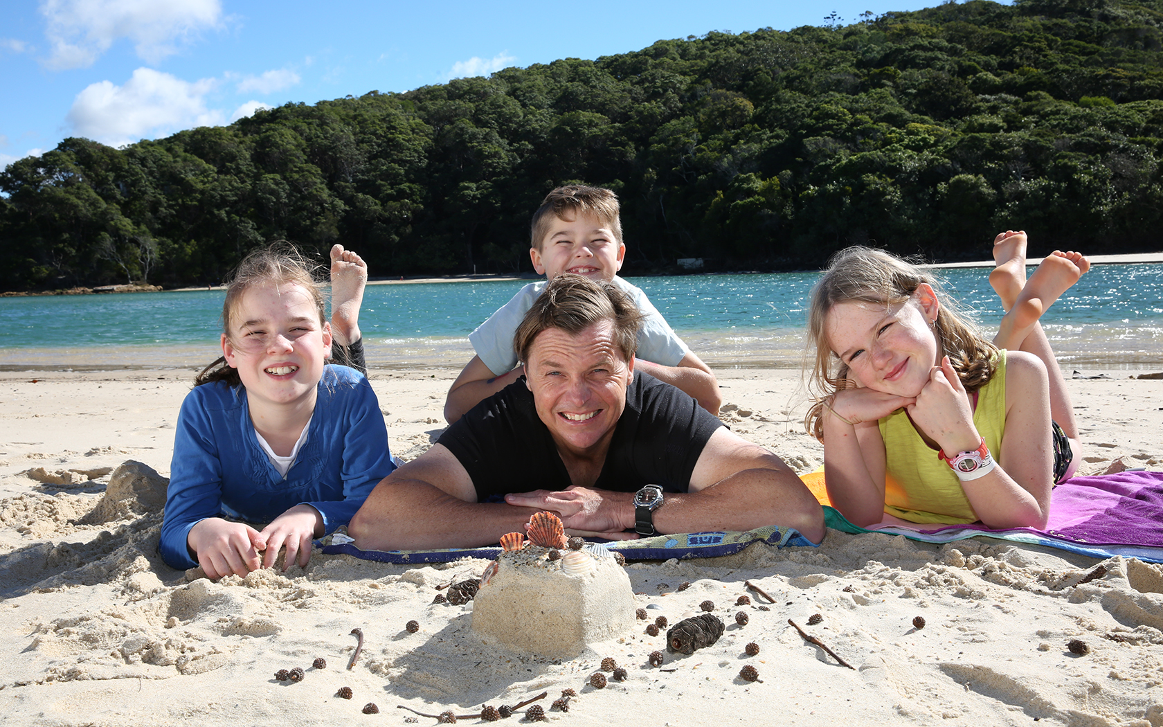 Family laying on the sand with water and trees behind.