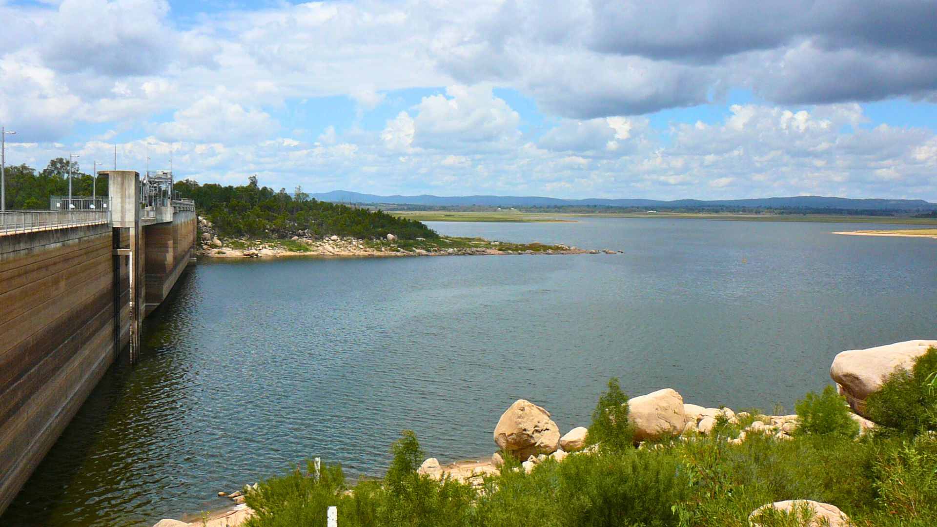 Dam wall and reservoir with rocky shoreline under a cloudy sky.