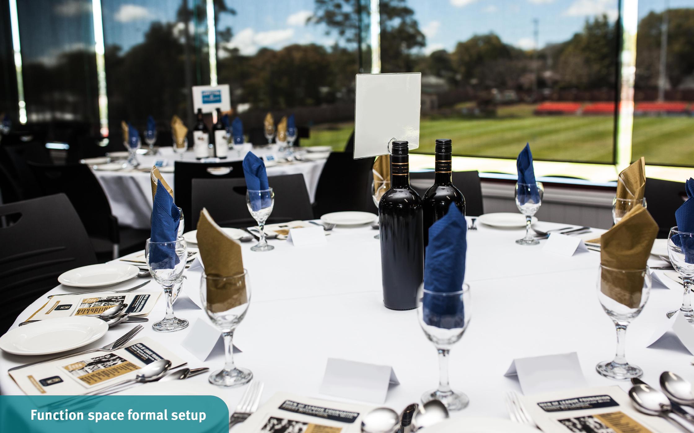 A round table with a white tablecloth, set for a group of people complete with plates and cutlery and two bottles of wine.