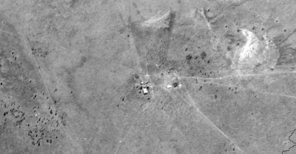 Black and white aerial photo of a grassland landscape. A small group of building is shown in the middle of the image, and there is very sparse vegetation shown.